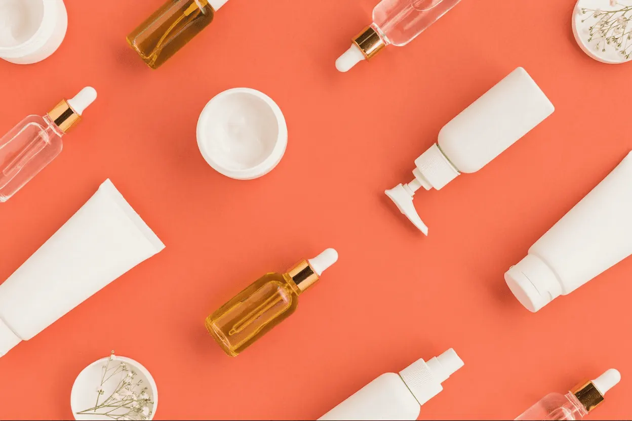 Skincare bottles, cosmetic jars, and serum droppers arranged on a coral background